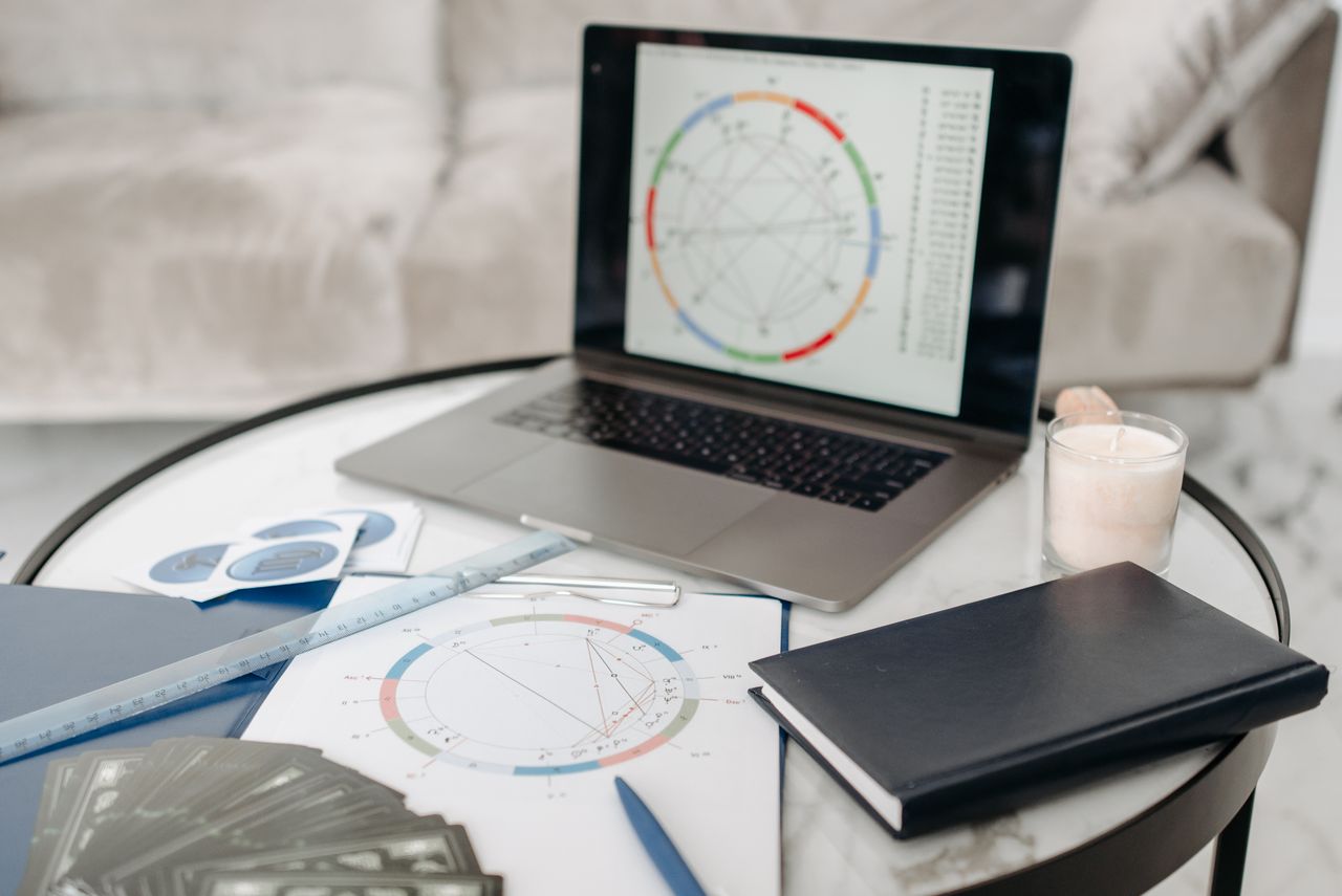 Astrology charts displayed on a laptop next to tarot cards and notebooks on a table.